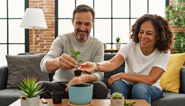 Middle-aged couple planting young seedling in sunlit living room pot, repotting together