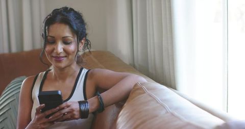 Woman relaxing on tan leather sofa scrolling smartphone by sunlit window, casual home moment