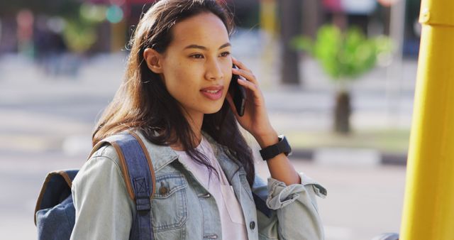 Young woman is using a smartphone while walking outdoors in an urban area. She is wearing a denim jacket and carrying a backpack, suggesting she may be a student or traveler. This image can be used in contexts related to communication, technology, lifestyle, urban living, travel, and everyday activities.