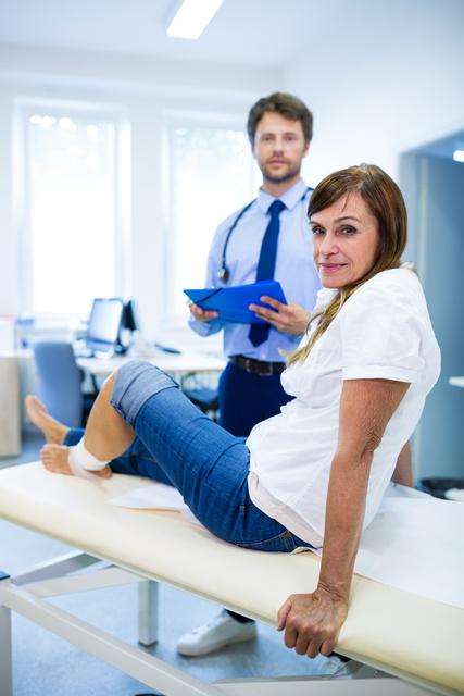 Patient sitting on examination table with leg bandaged, doctor holding clipboard in background. Ideal for healthcare, medical treatment, patient care, and hospital-related content.