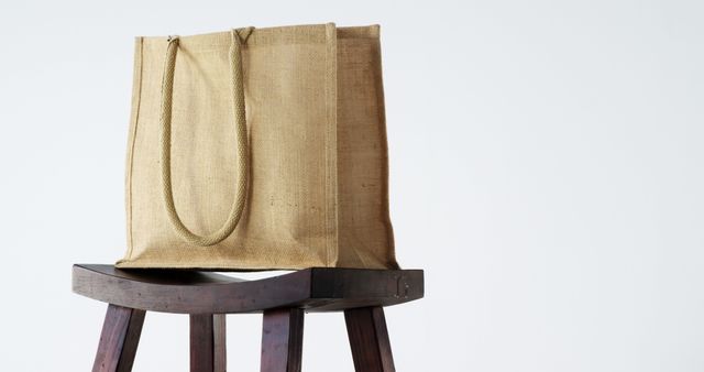 Eco-friendly jute tote bag placed on a wooden stool, captured against a plain white background. Ideal for themes related to sustainability, minimalism, and natural materials. Suitable for promoting environmental causes, reusable shopping bags, and minimalist living.