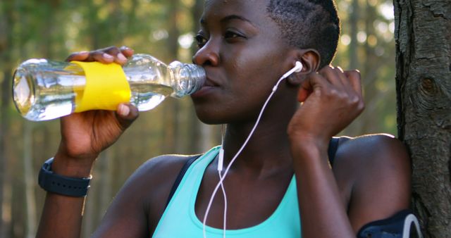 This visual represents a fit African American woman staying hydrated while exercising in a forest setting, emphasizing the importance of hydration and physical fitness. She is listening to music through headphones, suggesting an engaging and focused workout routine. This image is ideal for promoting health, fitness products, outdoor sports accessories, and lifestyle blogs.