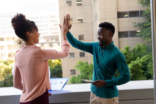 Business colleagues celebrating success with a high-five on an office balcony. Ideal for illustrating teamwork, collaboration, and corporate success in a professional setting. Can be used for business presentations, corporate websites, and promotional materials highlighting positive work culture.