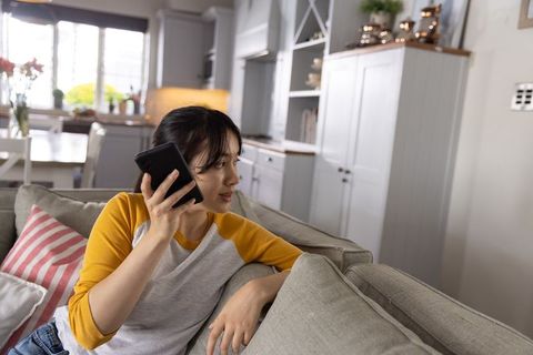 Asian woman talking on smartphone while relaxing on modern living room couch in casual home interior
