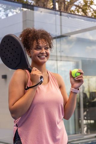 Smiling african american woman playing padel holding racket and ball on glass urban court