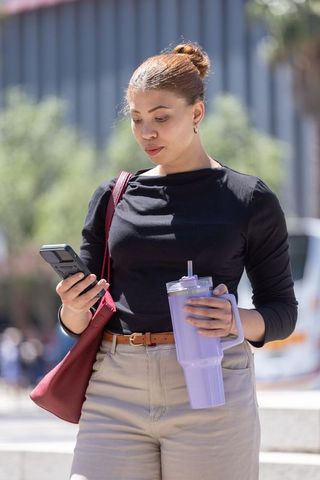 African american woman walking checking phone holding purple tumbler and red shoulder bag