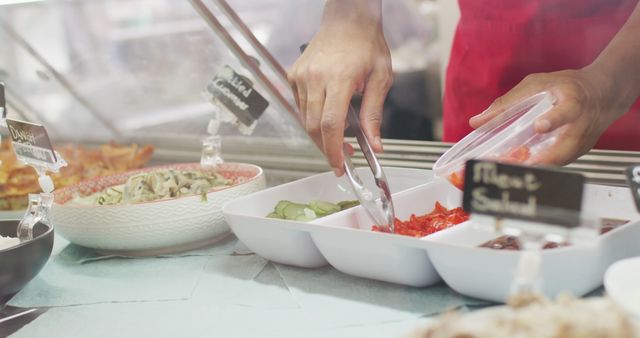 Person using tongs to prepare fresh salad at deli counter. Scene is associated with food preparation, nutrition, healthy eating. Suitable for use in content related to culinary arts, delis, food freshness, or healthy living.