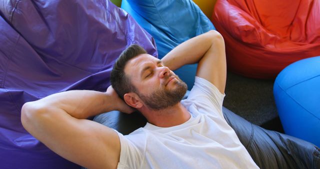 Man lying back and enjoying relaxation on colorful bean bags indoors. Perfect for use in lifestyle blogs, leisure-related content, or as part of promotional materials for relaxation and chilling products.