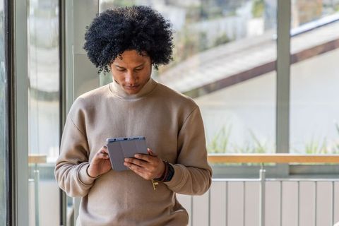 Young man using tablet in bright modern atrium wearing tan sweatshirt for remote work