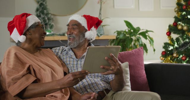 This image shows a happy senior couple wearing Santa hats, celebrating Christmas at home. They are smiling and using a tablet, possibly sharing holiday greetings or looking at photos. This image is perfect for promoting holiday products, greeting cards, social media posts about family togetherness, or advertisements for elderly services or technological products for seniors.