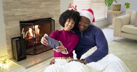Couple wearing Santa hat and reindeer antlers taking cozy selfie by roaring fireplace
