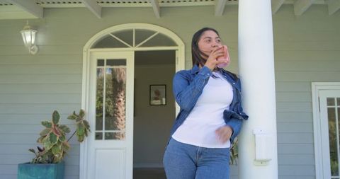 Woman sipping coffee from pink mug on sunny porch leaning against column in denim