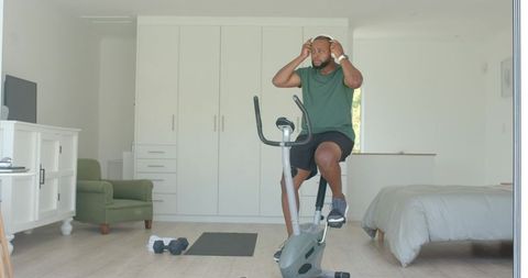 African american man adjusting headphones cycling on exercise bike in modern bedroom