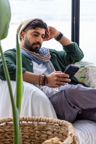 Relaxing man scrolling smartphone on sunlit sofa wearing beanie and scarf