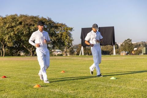 Diverse cricketers jogging and checking watches in training drill on grassy field