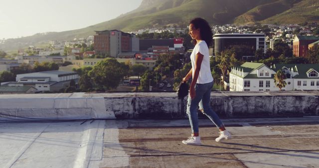Young woman walking on a rooftop with scenic urban background. The image shows city buildings set against a backdrop of lush hills and clear skies. Ideal for advertising urban lifestyle, freedom, exploration, casual fashion, and outdoor activities.