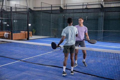 Diverse male players shaking hands at net on blue indoor padel court with glass walls and lounge are