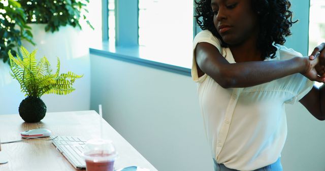 An African American woman appears to be stretching her arms while sitting at a modern workspace, with copy space. Her moment of self-care amidst a busy work environment emphasizes the importance of taking breaks for physical well-being.