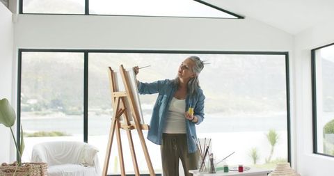 Senior woman painting at wooden easel in bright waterfront studio with large windows