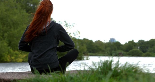 Back view of a woman with red hair meditating by a tranquil lake. Surrounded by lush green nature, she is practicing mindfulness and relaxation. Ideal for content on mental wellness, outdoor activities, relaxation techniques, and peaceful settings.