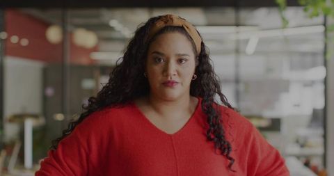 Confident woman standing in modern open office wearing red sweater and tan headband, facing camera