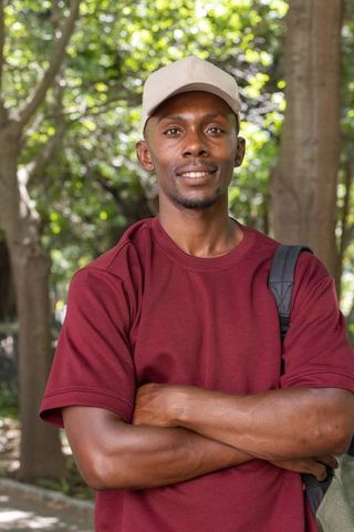 Confident african man standing in shaded park crossing arms wearing maroon tee and beige cap
