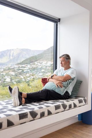 Mature man relaxing on window seat overlooking hillside in modern minimalist home with panoramic vie