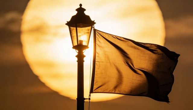 Golden backlit ornate street lamp and billowing flag at harbor with giant sun silhouette