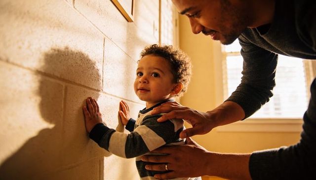 Sunlit father steadying toddler against wall during golden hour, tender parent-child bond
