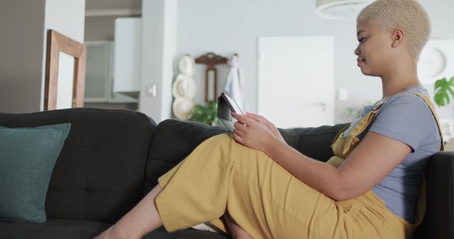 A young adult woman with short hair wearing yellow overalls sits on a dark comfortable couch in a modern home environment using a digital tablet. This could be used for promoting home automation technology, comfort in living spaces, online communications, or lifestyle content focused on relaxation and technology use at home.