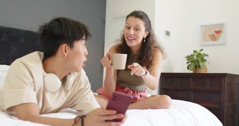 Intercultural couple chatting on bed holding mug and smartphone in cozy bedroom