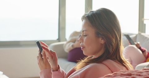 Woman Relaxing on Sofa Scrolling Smartphone While Friend Lounging in Sunlit Living Room