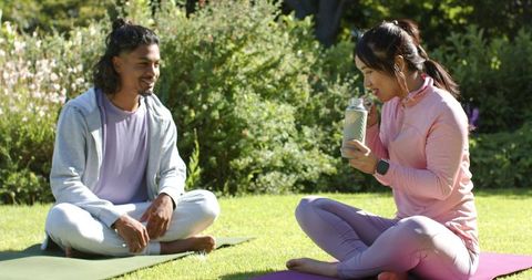 Indian man and asian woman practicing yoga outdoors, sharing water and relaxing in garden