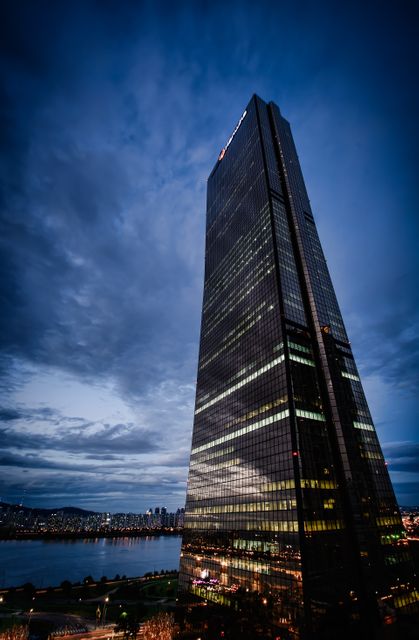 Featuring a towering skyscraper in the evening, equipped with reflective glass walls against a dusk sky. City lights start illuminating in the distance, catching early night atmosphere. This visual is suitable for representations about urban development, corporate business settings, architectural marvels, and modern city life.