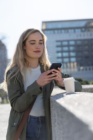 Young woman checking smartphone while enjoying takeaway coffee on urban plaza ledge