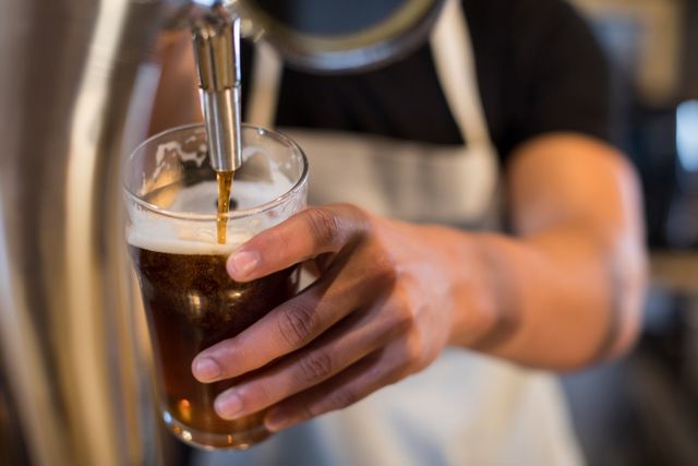 Midsection view of bartender pouring beer from tap into glass. Ideal for use in articles or advertisements related to bars, pubs, nightlife, hospitality industry, and alcoholic beverages. Can also be used for illustrating concepts of service, refreshment, and social gatherings.