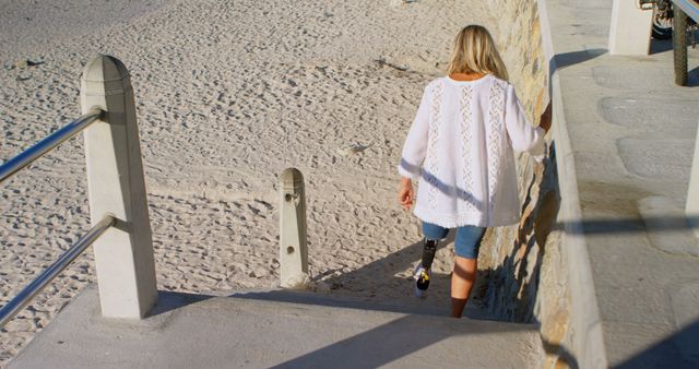 Woman with a prosthetic leg carefully walking down stairs to a sandy beach. Ideal for promoting inclusion, accessibility, summer activities, outdoor adventures, and stories of courage and independence.