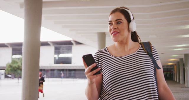 Smiling plus size caucasian woman wearing headphones and using smartphone walking in city street. City living, travel, communication and modern urban lifestyle.