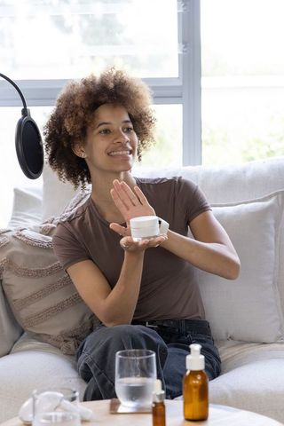 African american beauty creator demonstrating skincare cream on sofa in sunlit room