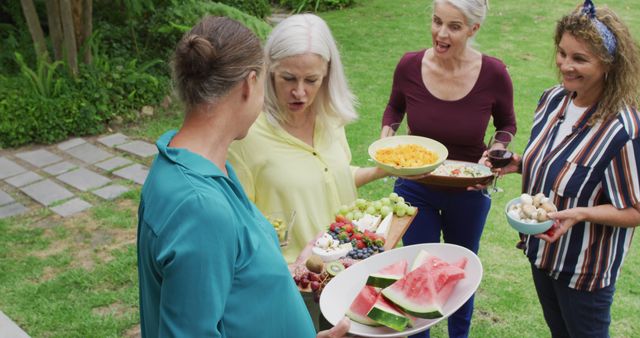 Senior friends are enjoying a summer garden party with fresh fruits and snacks. They are chatting and smiling while holding plates of food like watermelon, grapes, and other delicacies. The setting indicates a casual and happy occasion perfect for promoting socializing, healthy lifestyle, senior activities, and seasonal parties catered to older adults.