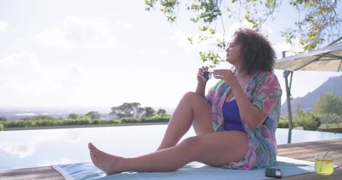 Curly woman relaxing at infinity pool wearing blue swimsuit and floral kimono