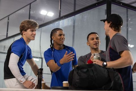 Multicultural male teammates chatting, laughing and hydrating at indoor court with gear