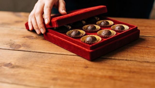 Hand lifting red velvet chocolate box revealing nine gourmet truffles on wood table
