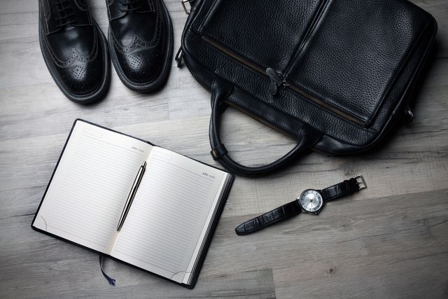 Collection showing business accessories arranged on wooden surface. Includes leather shoes, leather briefcase, blank open notebook with pen, and wristwatch. Useful for representing professionalism, business travel, or office essentials.