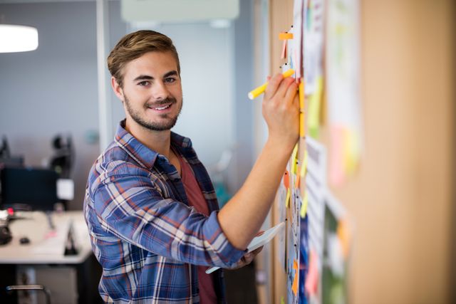Young man writing on sticky notes on a board in a modern office. Ideal for content related to business planning, creative brainstorming sessions, teamwork, project management, and office environments.