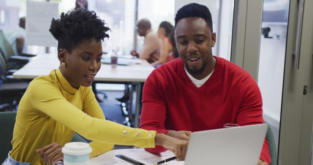 African American colleagues discussing a project and collaborating on a laptop in a modern office environment. Both are dressed in casual attire, sitting at a desk with notebooks and a coffee cup. Ideal for depicting teamwork, professional collaboration, modern workspaces, and project development scenarios.