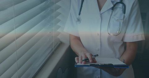 Nurse writing patient notes by window with stethoscope and clipboard in clinic