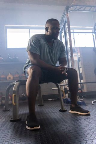 African man resting on bench in gym, clasping hands and focusing after strength training
