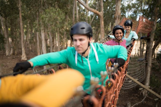 Group of friends walking on a rope bridge in a forest, wearing safety gear and helmets. Ideal for use in content related to outdoor activities, adventure sports, teamwork, and nature exploration.