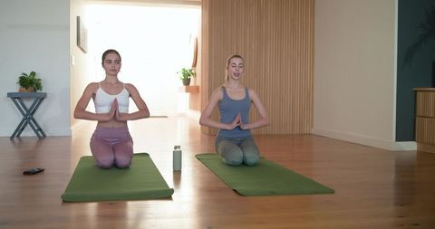 Two women practicing yoga kneeling prayer pose at home on green mats with water bottle